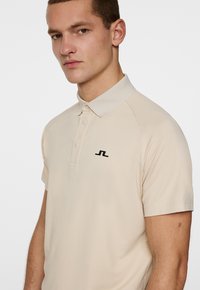 Beige pikétröja i mjukt tyg, med krage, korta ärmar och en svart logotyp på vänster bröst.