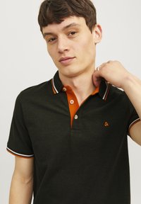 Donkergroen polo-shirt met een oranje kraag, witte bies op de mouwen en een klein geborduurd logo. Bevat twee knopen bij de hals.