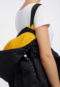 Czarna kurtka z żółtym polarem od środka, boczną kieszenią na zamek oraz sznurkiem z ozdobnym koralikiem. Nosi się ją na ramieniu, z widocznym paskiem naramiennym.