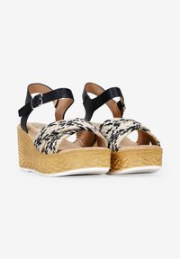 Tirantes de cuero negro en el tobillo y parte superior trenzada en beige y negro en sandalias de cuña con plataforma texturizada beige y suela blanca.