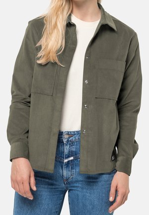 Kobieta ubrana w oliwkowo zieloną koszulę na guziki, na białym topie i niebieskich dżinsach, stojąca na jednolitym jasnym tle.