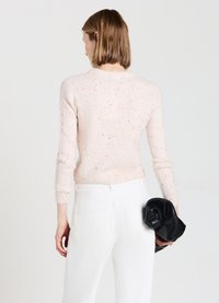 Helles rosa Strickpullover mit Sprenkeln, ausgestattet mit einem Rundhalsausschnitt und gerippten Manschetten, kombiniert mit weißen Hosen. Modell hält eine schwarze Tasche.