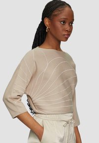 Beżowy sweter dzianinowy z białymi wzorami w paski, szerokim dekoltem w łódkę i rękawami o długości trzech czwartych, w połączeniu z jasnymi spodniami z troczkiem.