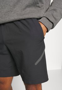 Pantalones cortos oscuros de color gris para hombre, con una textura suave, que cuentan con un bolsillo lateral y un acento reflectante en la pierna para mayor visibilidad. Se combinan con una sudadera gris.