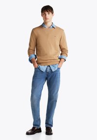 Un pulover tricotat de culoare maro deschis, suprapus peste o cămașă din denim albastru, asociat cu blugi albastre și mocasini maro închis. Contrast clar între design și culori.