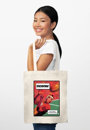 Mladá žena se usmívá a drží béžovou tašku přes rameno s vintage motivem fotbalového hráče a komiksovým nápisem „Touchdown!“