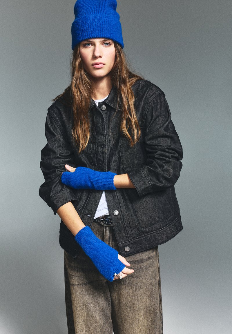 Schwarze Jeansjacke über einem weißen Hemd, kombiniert mit fingerlosen blauen Strickhandschuhen und einer blauen Mütze. Braune weite Hosen runden das Outfit ab.