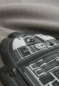 Grafika R2-D2 nadrukowana na szarym materiale, z czarnymi i białymi detalami, teksturowaną powierzchnią oraz charakterystycznymi okrągłymi i prostokątnymi kształtami.