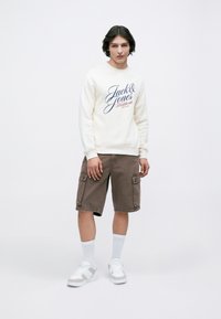 Mladý muž v bielom mikině s nápisom "Jack & Jones", hnedých cargo šortkách, bielych ponožkách a bielych teniskách, stojaci na pozadí bez vzoru.