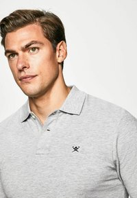 Grå polo-shirt med krave, prydet med et lille sort broderet logo på venstre bryst, lavet af tekstureret stof.