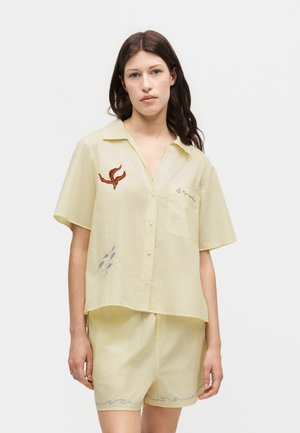 Ung kvinde med langt brunt hår har en løs, bleg gul kortærmet skjorte med abstrakt broderi og matchende shorts med bølgemotiver.