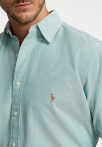 Uomo che indossa una camicia a maniche corte di colore verde chiaro con un piccolo logo ricamato di un giocatore di polo su cavallo sul petto, in primo piano sulla parte superiore del torso.