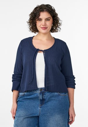Kvinde med krøllet hår iført en marineblå ribstrikket cardigan over en hvid top og blå højttaljede jeans, stående foran en hvid baggrund.