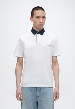 Jeune homme aux cheveux courts portant un polo blanc avec un col noir et un jean bleu clair, debout devant un fond blanc uni.