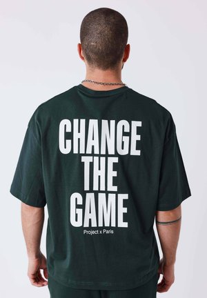 Homme portant un t-shirt noir avec texte blanc "CHANGE THE GAME" et "Project x Paris", vu de dos sur un fond uni.