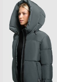 Ung kvinde iført en mørkegrå puffjakke med stor hætte over en sort top, der vender mod venstre foran en lys ensfarvet baggrund.