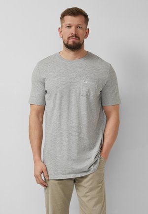 Mężczyzna z brodą, ubrany w szary T-shirt z krótkim rękawem i kieszenią na piersi oraz beżowe spodnie, stojący na tle jednolitego jasnoszarego tła.