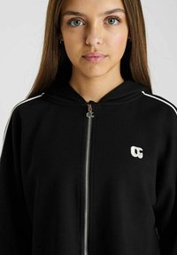 Zwarte zip-up hoodie met een wit logo, voorzien van een capuchon, twee-tone strepen op de mouwen en een gladde textuur. Zichtbare ritssluiting.