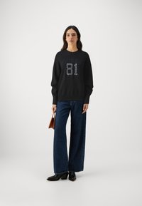 Svart sweatshirt med "GUESS 81" i silverpaljetter, matchad med vida mörk denimjeans och svarta klackboots. Håller en brun handväska.