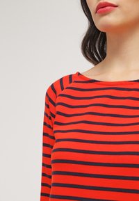 Haut rayé rouge et marine à manches longues avec un col large et une texture douce. Comprend des manches raglan et une coupe ajustée.