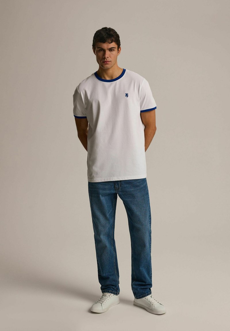 Biała koszulka z krótkim rękawem z niebieskimi wykończeniami, zestawiona z niebieskimi dżinsami i białymi trampkami. Koszulka ma małe logo na piersi.