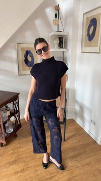 Γυναίκα που χαμογελάει σε εσωτερικό χώρο, φοράει μαύρο ριμπ αμάνικο τοπ, διάφανο παντελόνι με σχέδια σε ναυτικό μπλε, μαύρα μπαλαρίνια και μεγάλα μαύρα γυαλιά ηλίου.