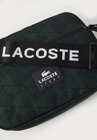 Borsa sportiva Lacoste verde a fantasia con tracolla nera e scritta bianca, tessuto testurizzato e logo brand rettangolare.