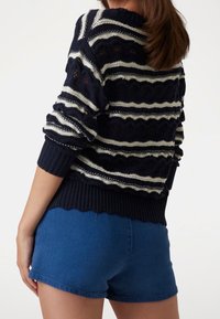 Il maglione lavorato a maglia presenta righe orizzontali blu navy e bianche con motivi a onda. Il maglione ha un collo e un orlo inferiore a costine. Abbinato a pantaloni corti blu.