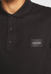 Musta polo-paita puuvillasta, jossa on kaulus, kolme nappia ja suorakulmainen etiketti, jossa on valkoisella teksti "Calvin Klein".
