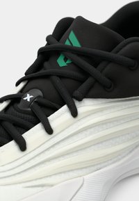 Black athletic shoe featuring textured materials, prominent white midsole with ridges, and a green logo detail near the laces.