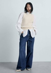 Gilet a coste beige indossato sopra una camicia bianca con colletto, abbinato a jeans larghi in denim blu e scarpe beige a punta, il tutto su uno sfondo chiaro.