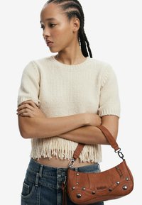 Brun lædertaske med dekorativ syning og metal accenter, parret med en cremefarvet sweater med frynser og mørkeblå denim jeans.