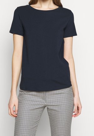 Kobieta ubrana w gładki, ciemnogranatowy t-shirt z krótkim rękawem oraz spodnie w czarno-biały wzór w jodełkę, stojąca na jednolitym tle.
