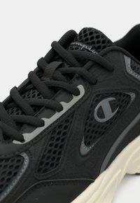 Κοντινό πλάνο μαύρου αθλητικού παπουτσιού με πλέγμα και συνθετικές επιφάνειες, μαύρα κορδόνια και γκρι λογότυπο Champion στο πλάι, λευκή σόλα ορατή.