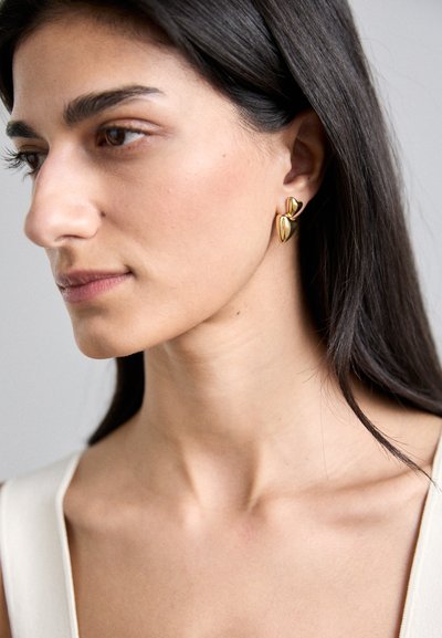 Boucles d'oreilles dorées avec une forme abstraite comprenant deux formes enroulées semblables à des feuilles se chevauchant. Surface lisse avec une finition brillante.