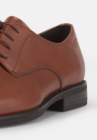 Chaussure en cuir brun avec un bout arrondi, un laçage et un petit talon en bois noir. Texture lisse avec des détails de couture subtils.