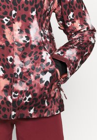 Gros plan d'une personne portant une veste rouge et noire à motif léopard, main dans la poche latérale, associée à un pantalon rouge foncé uni.