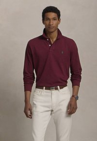 Bordo dolginasleka polo majica s kragnom, z majhnim zelenim logotipom. V kombinaciji z belimi kavbojkami in rjavim pasom. Model nosi ura.
