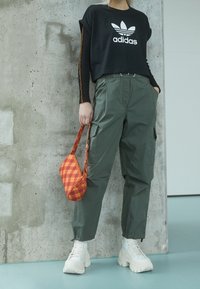 Person wearing black Adidas cropped top, olive cargo pants, white platform boots, holding orange checkered handbag, standing by concrete wall.