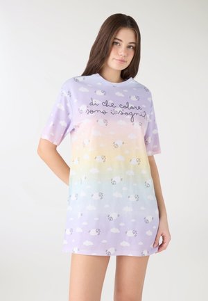 Giovane donna che indossa una camicia da notte a gradiente pastello con nuvole, motivi di pecore e testo italiano sul petto, in piedi con le mani lungo i fianchi.