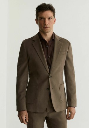 Młody mężczyzna stojący, ubrany w brązową marynarkę i dopasowane spodnie, pod którymi ma ciemnobrązową koszulę na guziki, na neutralnym tle.