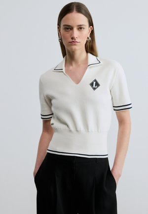 Jonge vrouw die een wit gebreid topje met korte mouwen en zwarte bies, zwarte cropped broek en zilveren oorringen draagt, staand met handen in de zakken.