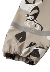 Tissu beige résistant à l'eau avec des motifs animaliers abstraits en noir et blanc. Détail de poignet froncé avec une bande d'accent en argent portant un logo.
