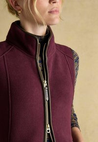 Bordeauxfarbener Fleece-Weste mit hohem Kragen, goldigem Reißverschluss und braunen Akzenten. Darunter ist ein langärmliges Oberteil mit blauem Muster sichtbar.