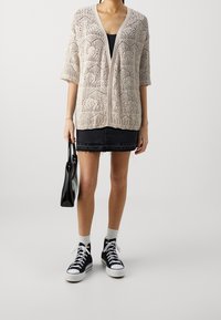 Béžový pletený kardigan s otevřeným předním dílem, krátkými rukávy a složitým krajkovým vzorem, doplněn�ý o černou denimovou minisukni a tenisky.
