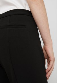 Pantalon noir avec une texture lisse, présentant une poche arrière dissimulée. Des lignes épurées et une coupe ajustée mettent en valeur le design minimaliste.