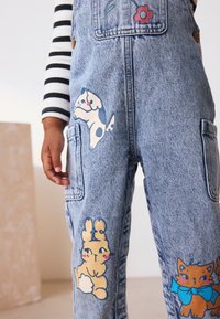Denim-overalls har blåt stof med legesyge dyreillustrationer: en hund, en kanin og en kat. Designet inkluderer lommer og livlige farver.