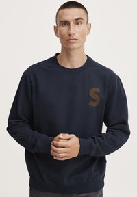Hanorac albastru marin din material moale, cu guler rotund, maneci lungi și un logo aurie "S" pe piept. Croiala regulară și manșete ribbate.
