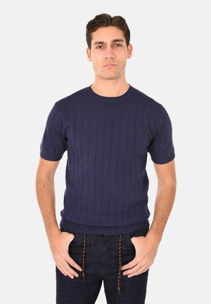 Uomo che indossa un maglione a maniche corte a coste blu navy e jeans blu scuro con le mani nelle tasche, in piedi davanti a uno sfondo chiaro uniforme.