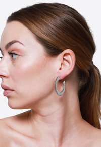 Orecchini a cerchio argento con una superficie texturizzata, forma circolare e un design aperto, indossati da una persona con un incarnato liscio.